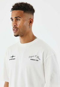 Joven con corte de pelo rizado y desvanecido, vistiendo una camiseta blanca de “Project X Paris Members' Club”, collar de cadena plateada, anillos en la nariz y un pendiente de cruz, mirando hacia la izquierda.