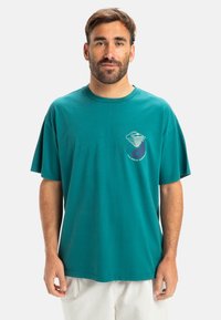 Camiseta de algodón en color verde azulado con escote redondo y mangas cortas; presenta un diseño gráfico en blanco y lavanda en el pecho.