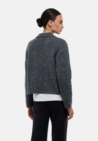 Veste courte en mélange de laine grise, dotée d'un col revers et de manches longues. Tissu texturé avec un motif subtil, associé à une chemise blanche.