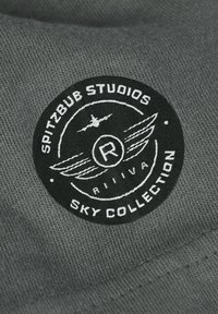 Ronde zwarte patch met witte tekst "Spitzbub Studios Sky Collection," een gevleugeld embleem en een kleine vliegtuigillustratie op grijze stof.