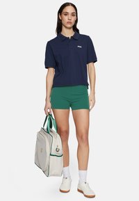 Marineblå pikétröja med en logotyp, gröna shorts, vita sneakers och en grå väska med gröna accenter och dragkedjedetaljer.