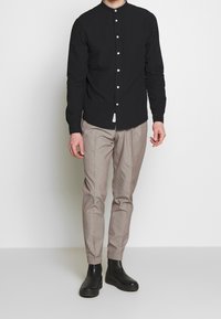 Czarna koszula z długim rękawem, stojącym kołnierzem i zapięciem na guziki z przodu, zestawiona z jasnobrązowymi, wzorzystymi spodniami i czarnymi botkami za kostkę.