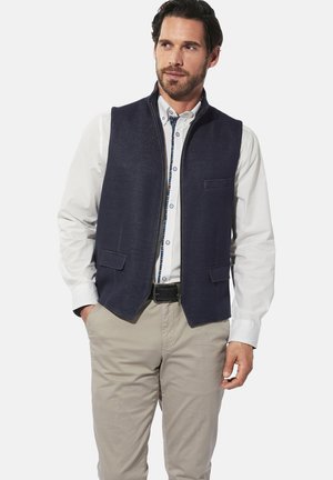 Mann in weißem Hemd mit Knopfleiste, marineblauer Zip-Weste, beigen Hosen und schwarzem Gürtel, der mit Händen in den Taschen vor einem weißen Hintergrund steht.