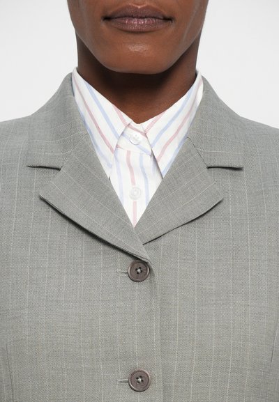 Svetlošedý blazer s prúžkami a zúženým golierom, so zlatým zapínaním na dva hnedé gombíky, položený na bielej košeli s červenými a modrými prúžkami. Textúrovaná látka.