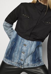 Černá džínová bunda s dlouhými rukávy, ozdobená stříbrnými hroty, s knoflíkovým zapínáním a dvěma náprsními kapsami. Džínová část má poškozený vzhled.