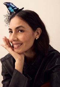 Zwarte leren jas, blauw haaraccessoire met pailletten en tule, druppelvormige oorbellen, kort donker haar, glimlachend, hand onder de kin.