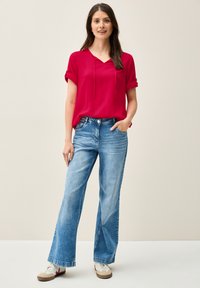 Blusa rossa a maniche corte con coulisse e risvolti, abbinata a jeans ampi blu chiaro e sneakers bianche con dettagli rossi.