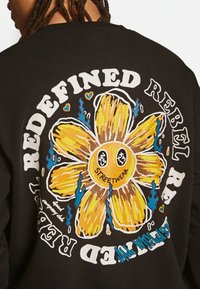 Schwarzer Hoodie mit einer großen gelben Blumen-Grafik mit ausdrucksvollen Gesichtszügen, blauen Flammen und dem Text "STREETWEAR" und "REDEFINED REBEL."
