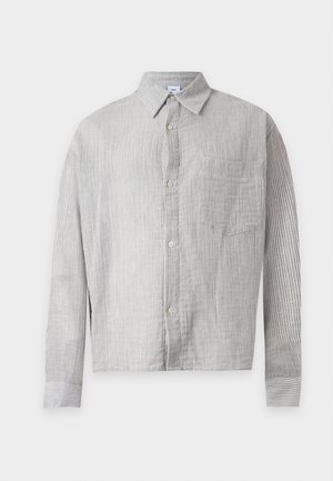 Joven de pie con una camiseta gris de manga larga, pantalones cortos vaqueros azules claros hasta la rodilla, calcetines negros y zapatos negros sobre un fondo blanco.