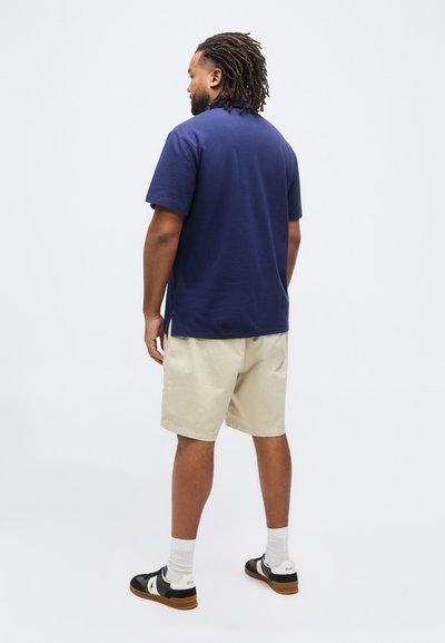 Άνδρας με κοτσίδες, ντυμένος με μπλε πόλο πουκάμισο, μπεζ σορτς, άσπρες κάλτσες και μαύρα-άσπρα αθλητικά παπούτσια, στέκεται μπροστά σε απλό φόντο.