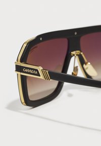 Gafas de sol negras de gran tamaño con lentes marrón degradado, detalles en oro en la montura y logo "CARRERA" en el lateral. Detalle texturado en las patillas.