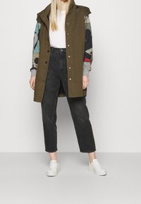 Olijf groene parka met gemusterde mouwen, gecombineerd met hoogzitende zwarte jeans en witte sneakers. Voorzien van drukknoopsluiting en capuchon.