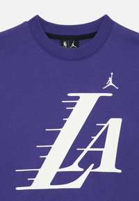 Camiseta de algodón púrpura con un gran gráfico blanco de "LA" y un logotipo de Jumpman. La camiseta tiene un cuello redondo clásico y mangas cortas.