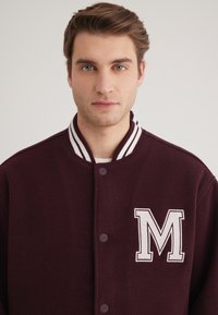 Burgunderfarbene Collegejacke aus strukturierter Stoff mit weißem Rippkragen, versehen mit einem gestickten weißen "M" auf der linken Brust und Knopfverschluss.