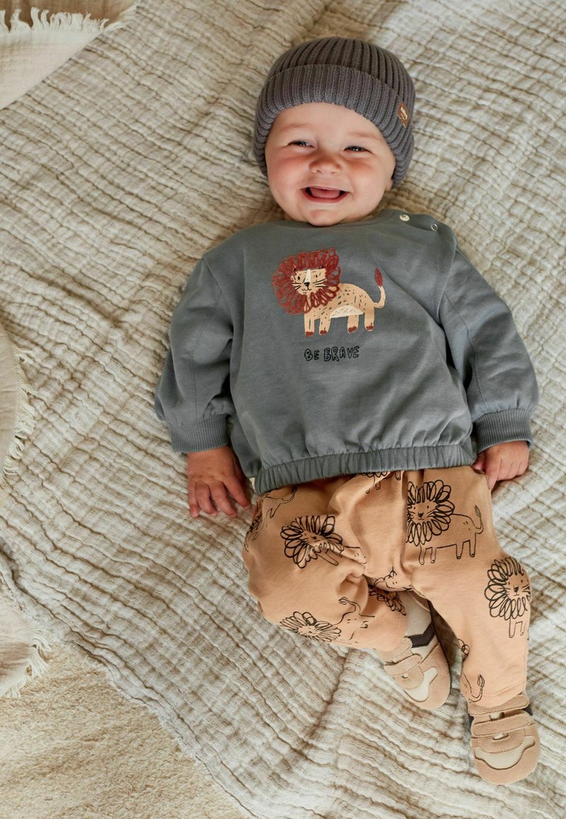 Grå sweatshirt med løveprint og teksten "Vær Modig," parret med lysebrune bukser med løvemønster. Blødt strikhue. Ligger på en struktureret tæppe.