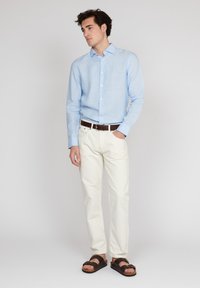 Chemise bleu clair à manches longues, pantalon blanc et sandales marron. La chemise a un col classique et un devant à boutons. Détails de ceinture marron.