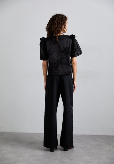 Juodas viršus su pūstomis rankovėmis, su sudėtingu karoliukų ir gėlių siuvinėjimu. Pora su plačiomis juodomis kelnėmis. Lygi audinio tekstūra.