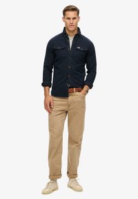 Uomo in piedi che indossa una camicia button-up blu navy, pantaloni beige con una cintura marrone, sneaker beige e un orologio, contro uno sfondo bianco.