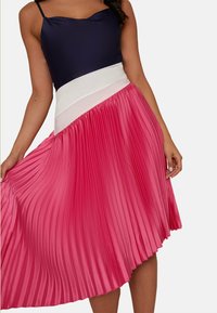 Faltrock mit einem pinkfarbenen unteren Abschnitt und einem weißen Bund, kombiniert mit einem dunkelblauen Oberteil mit gewelltem Ausschnitt.