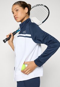 Jovem mulher com um casaco desportivo azul-marinho e branco, segurando uma raquete de ténis sobre o ombro e a colocar uma bola de ténis no bolso do casaco.