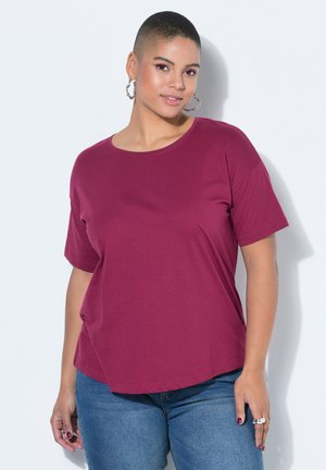Woman with short hair wearing hoop earrings, a magenta short-sleeve shirt, and blue jeans, standing against a white wall.