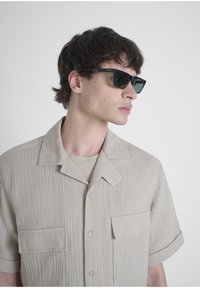 Camicia beige a maniche corte con texture, due tasche sul petto, colletto a picco e chiusura a bottoni, abbinata a occhiali da sole quadrati neri.