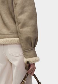 Bruine leren jas met sheepskin afwerking aan de manchetten, met een volumineus mouwontwerp. Lichtgekleurde broek en een handtas zijn gedeeltelijk zichtbaar.