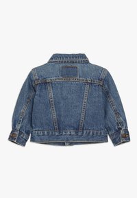 Chaqueta de mezclilla en azul medio con un cuello clásico, puños abotonados y detalle de yugo en la parte trasera. Presenta costuras cosidas y un parche cuadrado en la parte posterior.