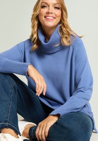 Pull à col roulé bleu clair en tissu maille douce avec poignets et encolure côtelés, associé à un jean en denim foncé et des baskets blanches.