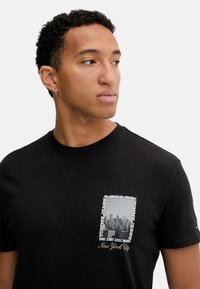Camiseta de algodón negra con un bolsillo en el pecho que presenta un gráfico de un horizonte urbano, el texto "Nueva York" y acentos de marca.