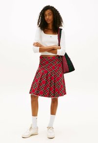Mujer de pie con los brazos cruzados, llevando una blusa blanca de manga larga, una falda de cuadros rojos, calcetas blancas, zapatillas blancas y llevando un bolso de cuadros.