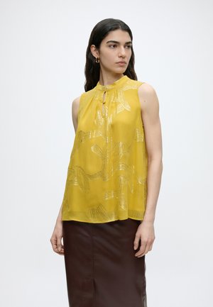 Mujer de cabello oscuro lleva blusa sin mangas amarilla mostaza con patrón abstracto y falda marrón, mirando de lado contra un fondo liso.