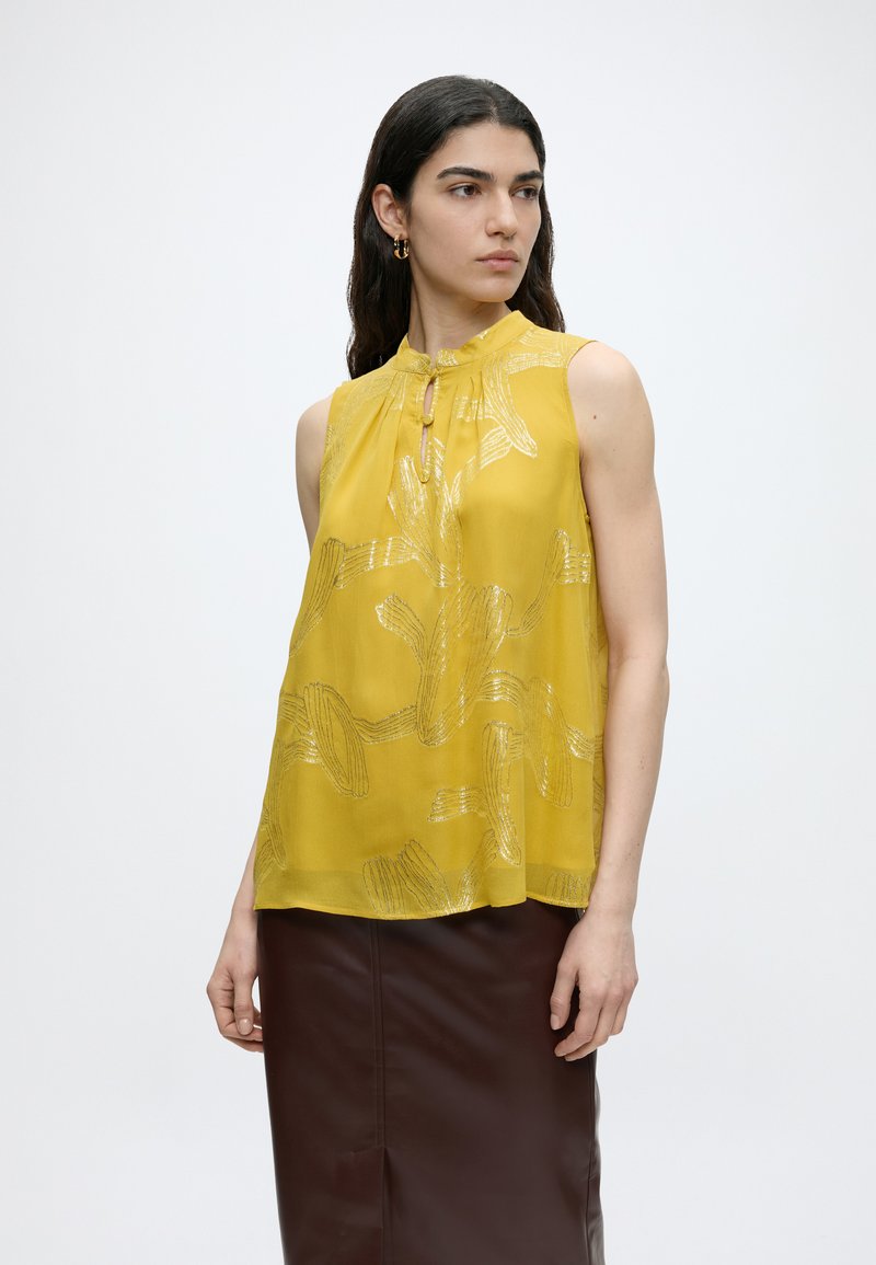 Γυναίκα με σκούρα μαλλιά φοράει αμάνικη μουσταρδί μπλούζα με αφηρημένο σχέδιο και καφέ φούστα, κοιτάζει πλάγια σε απλό φόντο.