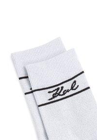 Chaussettes côtelées gris clair avec une bande noire horizontale et un logo script en haut. Texture douce avec une forme de chaussette standard.