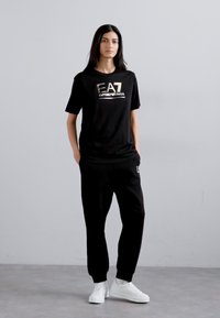 Zwarte katoenen T-shirt met metallic logo, gecombineerd met bijpassende zwarte joggers. Model in witte sneakers staat tegen een neutrale achtergrond.