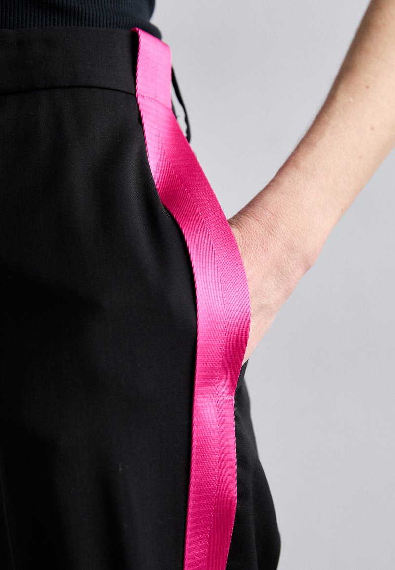 I pantaloni neri presentano un'evidente nastro di raso rosa brillante lungo il lato. Una mano è posata nella tasca, evidenziando il design e la texture.