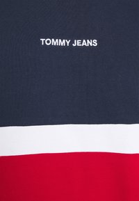Námorně modré, bílé a červené látkové pruhy s nápisem "TOMMY JEANS" vyšitým bílou barvou uprostřed modré části.