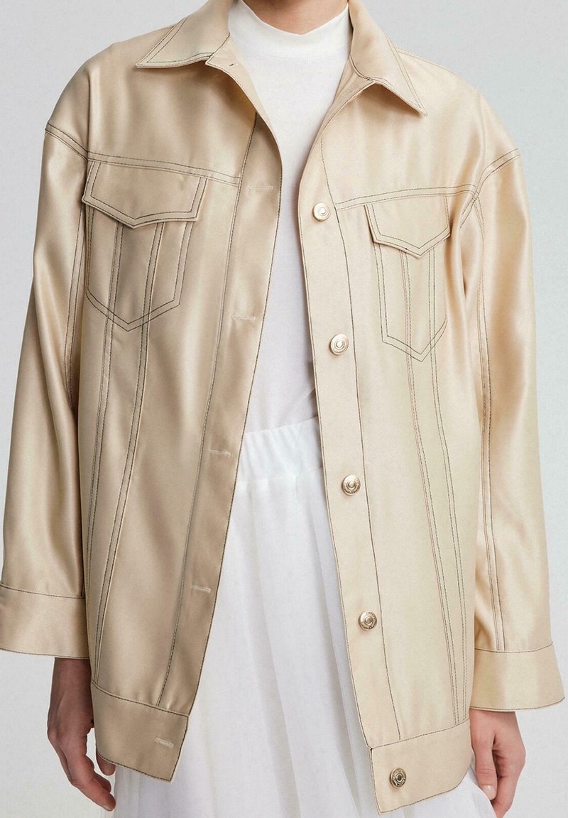 Veste en satin beige avec deux poches poitrine, fermeture à boutons simple boutonnage, surpiqûres contrastées et manches longues. Texture lisse.