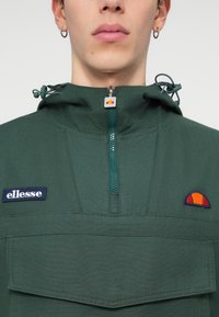 Zielony lekki pulower z półzamkiem, z logo naszywką, regulowanym ściągaczem w kapturze oraz detalem kieszeni z przodu.