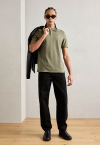 Un tricou polo verde măslin cu mâneci scurte, purtat cu pantaloni negri cu croială largă. O geacă de piele neagră este aruncată pe un umăr. Ochelari de soare negri.
