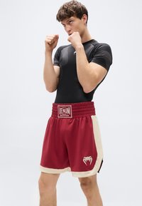 Μαύρο εφαρμοστό αθλητικό μπλουζάκι και κόκκινο σορτς με κρεμ λεπτομέρειες. Το σορτς διαθέτει το λογότυπο Venum στη μέση και στο πλάι, με λείο υλικό συνολικά.