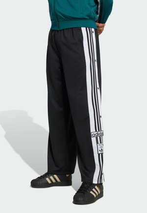 Osoba koja nosi crne Adidas trenirke s bijelim prugama i gumbima uz strane, uparene s crnim Adidas tenisicama s bež prugama.