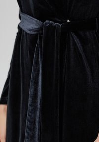 Robe en velours noir avec une texture douce, dotée d'une large ceinture nouée à la taille et d'un léger éclat, mettant en valeur le luxe et le confort.