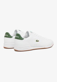 Witte leren sneakers met groene accenten, voorzien van een vetersluiting, zijkant perforaties en een bruine rubberen zool. Lacoste-logo op de hiel.