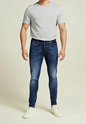Mand iført lysegrå kortærmet skjorte, smalle mørkeblå jeans og hvide slip-on sko, der står mod en ensfarvet baggrund.