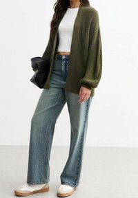 Oliivinvihreä neulottu neuletakki leveillä hihoilla, yhdistetty korkeavyötäröisiin vaaleansinisiin farkkuihin ja beigenvärisiin lenkkareihin. Pitelee mustaa clutch-laukkua.