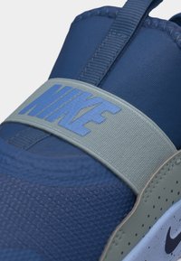 Modrá a sivá teniska Nike v detailnom zábere, zobrazujúca textilný zvršok s textúrou a remienok s veľkým logom Nike cez hornú časť.