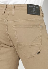 Pantalones de mezclilla beige resistentes con corte recto, bolsillos traseros cosidos y una etiqueta de tela en la cintura. Textura suave.