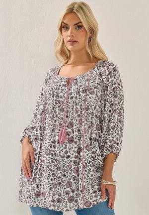 Donna bionda che indossa una camicetta larga con motivo floreale, accenti rosa e nappine, abbinata a jeans blu e braccialetti d'argento.