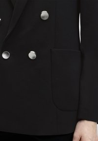Μαύρο διπλόμπουτο σακάκι με λείο υφή, διακοσμημένο με ασημί κουμπιά και τσέπη καpatch στη αριστερή πλευρά.